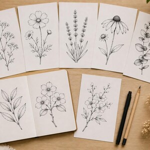 Puede incluir: Ilustraciones de arte lineal bot&aacute;nico de flores y follaje en tinta negra sobre papel blanco. La imagen incluye un cuaderno de espiral y tarjetas individuales. Tambi&eacute;n hay un l&aacute;piz y un bol&iacute;grafo.