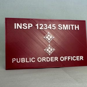 May include: A red plastic name tag with white text that reads "INSP 12345 SMITH PUBLIC ORDER OFFICER". The tag has two white star-shaped symbols.
