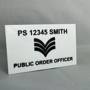 May include: White rectangular sign with black text that reads "PS 12345 SMITH" and "PUBLIC ORDER OFFICER". A black chevron symbol is centred above the text.
