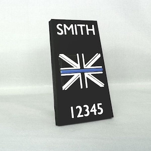 May include: Black rectangular sign with the name "SMITH" and the number "12345" in white. The sign features a white and blue graphic of the Union Jack flag with a blue line through the center.