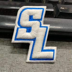 May include: White felt embroidered patch with the letters "SL" in blue outline. The patch is on a grey textured surface.