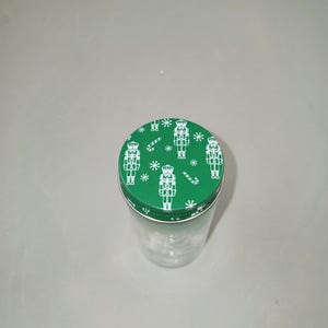 May include: A clear glass jar with a green lid featuring a repeating pattern of white nutcracker figures, snowflakes, and candy canes. The jar is empty and sits on a light gray surface. The lid is round and fits snugly on the jar.