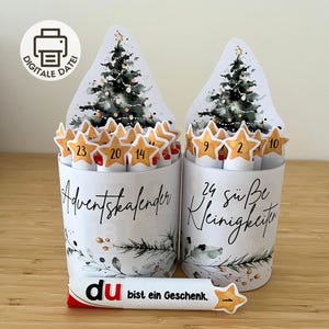Könnte beinhalten: Zwei zylindrische Behälter mit Aquarell-Weihnachtsbaum-Illustrationen und goldenen sternförmigen Anhängern, nummeriert von 1 bis 24. Die Behälter tragen den Text Adventskalender und 24 süße Kleinigkeiten. Eine einzelne Rolle zeigt den Text du bist ein Geschenk.