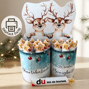 Pode incluir: Um conjunto de calendário do advento DIY com dois recipientes cilíndricos com uma cena de inverno em aguarela de dois veados. Os recipientes contêm tubos com estrelas numeradas de 1 a 24. O texto diz Adventskalender, 24 süße Kleinigkeiten e du bist ein Geschenk.