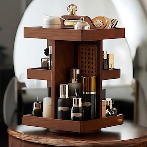 Könnte beinhalten: Ein dreistufiger, drehbarer Kosmetik-Organizer aus Holz. Der Organizer ist aus dunkelbraunem Holz gefertigt und verfügt über quadratische Regale. Auf den Regalen werden verschiedene Kosmetikprodukte, darunter Flaschen, Gläser und Puderdosen, ausgestellt. Ein Griff befindet sich auf dem obersten Regal.