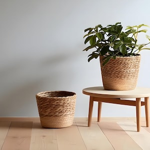 May include: Two woven baskets, one empty and one holding a green plant, sit on a light wood floor. A small, round wooden table with a plant in a woven basket is to the right. The baskets and table have a natural, neutral color palette.