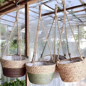 Puede incluir: Cuatro cestas colgantes tejidas con colgadores de cuerda. Las cestas son de varios tonos de marrón, beige y verde. Cada cesta tiene un forro de plástico blanco. Las cestas están suspendidas de ganchos metálicos.