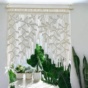 May include: Cream-colored macrame curtain with a leaf pattern, hanging in front of a window. The curtain is made of knotted cords, creating a textured design. The window sill holds potted plants with green leaves.