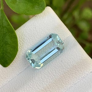 Puede incluir: Una gema rectangular de color azul claro con un corte facetado. La gema se muestra sobre una superficie blanca, con follaje verde en el fondo. Las facetas de la gema reflejan la luz, creando un efecto brillante. La gema es de color azul claro y transparente.