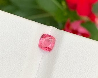Op de afbeelding: Een vierkant geslepen, roze edelsteen wordt getoond op een wit oppervlak. De edelsteen heeft een levendige roze tint, met een licht afgeronde vierkante vorm. De achtergrond toont groen gebladerte en rode bloemen.