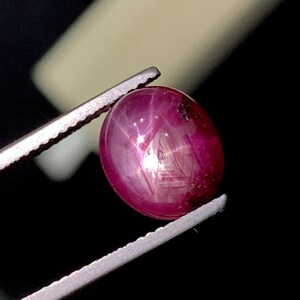 May include: A deep magenta oval-shaped star ruby gemstone with a smooth cabochon cut, displaying a distinct six-rayed star effect across its surface. The gemstone is held by a pair of metallic tweezers, showcasing its translucent and reflective quality.