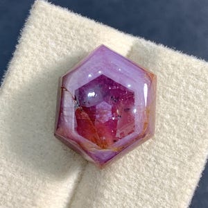 以下が含まれることがあります： 鮮やかなマゼンタと深い紫色のカラーパレットが特徴の六角形カットの宝石。石の表面はファセットカットされており、内部のインクルージョンや自然な筋模様が見え、幾何学的な形状全体に光を反射する磨き上げられた仕上げが施されています。