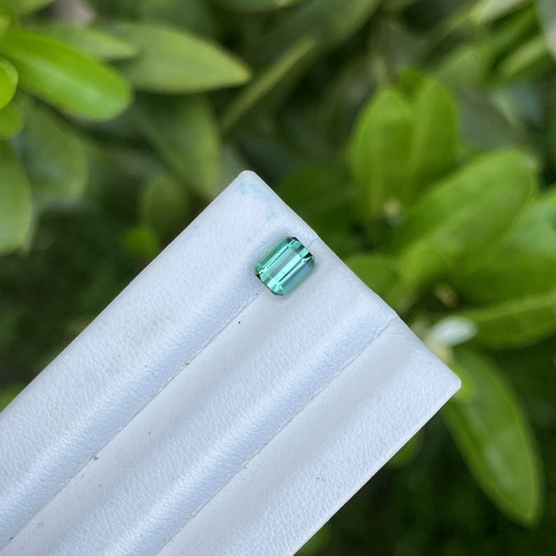 May include: A rectangular, teal-colored gemstone with a polished surface. The gem is set on a white, textured surface, with a blurred green foliage background. The gem is likely a tourmaline or similar precious stone.