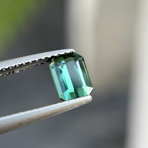 May include: A rectangular, emerald-cut gemstone with a teal hue is held by a pair of silver tweezers. The gem's facets reflect light, showcasing its clarity and color. The background is blurred, emphasizing the gem's details.