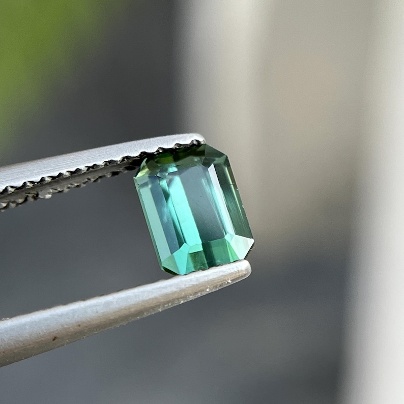 May include: A rectangular, emerald-cut gemstone with a teal hue is held by a pair of silver tweezers. The gem's facets reflect light, showcasing its clarity and color. The background is blurred, emphasizing the gem's details.