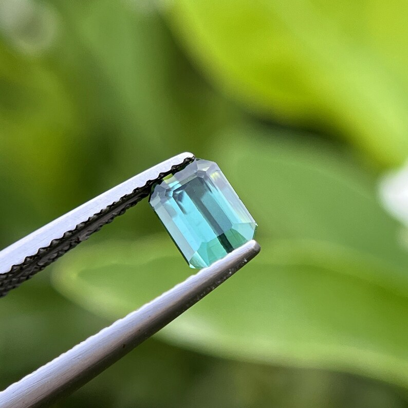 May include: A rectangular, teal-colored gemstone held by silver tweezers. The gem has a faceted cut, reflecting light and showcasing its color variations. The background is a soft, blurred green, providing a natural contrast to the gem's vibrant hue.