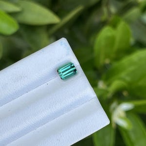 May include: A rectangular, teal-colored gemstone with a polished surface. The gem is set on a white surface, with a blurred green foliage background. The gem's facets reflect light, showcasing its clarity and color.