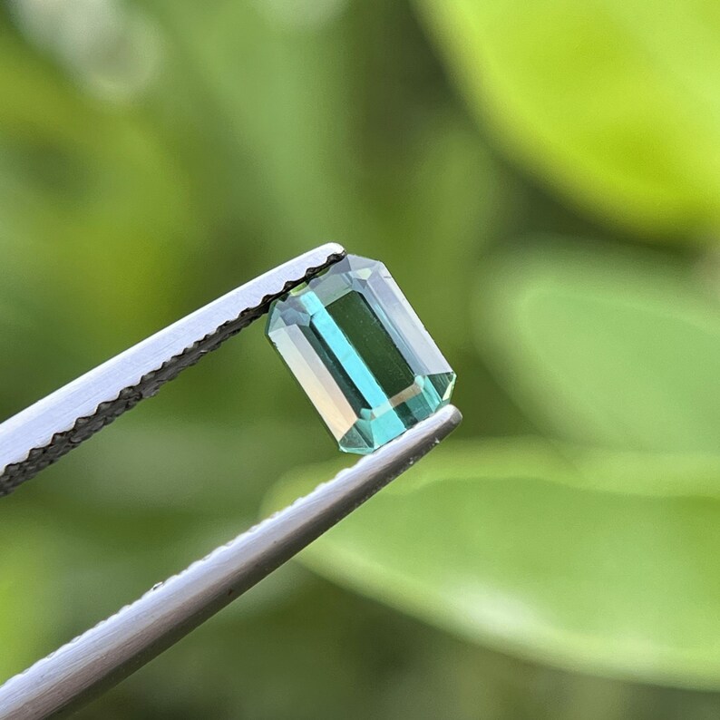 May include: A rectangular, emerald-cut gemstone with a teal and gold color gradient is held by a pair of silver tweezers. The gem is displayed against a blurred green background, highlighting its facets and clarity.