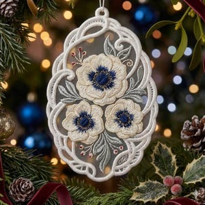May include: An oval-shaped white lace ornament featuring an intricate embroidered design of three cream-colored flowers with dark blue centers, surrounded by light green leaves and small red berries, all set against a sheer mesh fabric background.