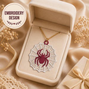 May include: A gold chain necklace featuring a circular pendant with a white embroidered spider web and a dark red embroidered spider at the center. The pendant is displayed inside a beige jewelry box. The text EMBROIDERY DESIGN is visible.