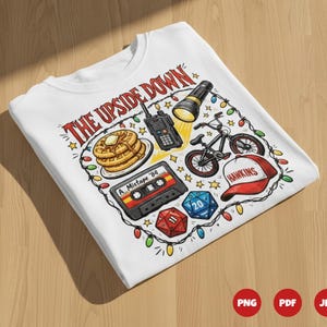 Puede incluir: Camiseta blanca con un diseño gráfico colorido con el texto "THE UPSIDE DOWN". El diseño incluye gofres, una cinta de cassette, un walkie-talkie, una bicicleta, una linterna, una gorra de béisbol y dados. La camiseta está decorada con luces de cadena.