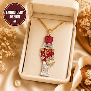 Op de afbeelding: Een goudkleurige ketting met een geborduurde notenkrakerhanger. De notenkraker draagt een rood jasje met gouden accenten, een rode hoed en een witte broek, en houdt een gouden trommel vast. De hanger wordt gepresenteerd in een beige doosje.