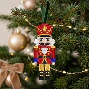 May include: A nutcracker ornament featuring detailed embroidery in red, gold, blue, and black thread. The figure wears a tall red hat, a red jacket with gold epaulets, and black boots. It hangs from a dark green velvet ribbon loop.