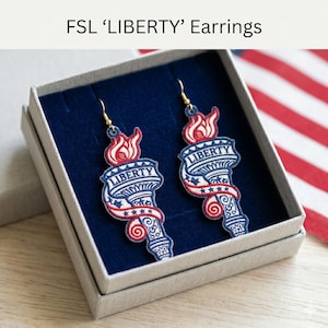 以下が含まれることがあります： ギフトボックスに入った「LIBERTY」イヤリングのペア。イヤリングは、赤、白、青の刺繍が施されたトーチのデザインが特徴です。「LIBERTY」の文字がトーチに表示されています。ゴールドカラーのフックが付いており、青色の内装のグレーの箱に入っています。