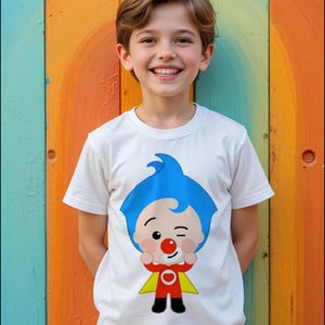 Może przedstawiać: Biały t-shirt z kr&oacute;tkim rękawem z nadrukiem graficznym przedstawiającym postać z kresk&oacute;wki z niebieskimi włosami, czerwonym nosem oraz w czerwonym stroju z ż&oacute;łtą peleryną. Postać mruga okiem i trzyma dłonie uniesione blisko twarzy na środku.