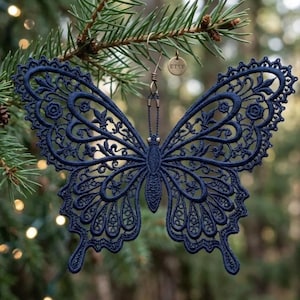 May include: A dark navy blue butterfly ornament crafted from intricate lace embroidery. The butterfly features detailed filigree patterns throughout its wings and body. A small circular metal tag with the text ETSY is attached to the top hook.