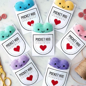 Pode incluir: Uma coleção de abraços de bolso em forma de coração feitos à mão, de crochê, em várias cores, incluindo azul, amarelo, verde, rosa e roxo. Cada coração está dentro de um cartão branco com o texto "POCKET HUG HANDMADE" e um coração vermelho. Botões, tesouras e barbante também estão presentes.