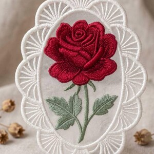 May include: An oval-shaped white lace ornament featuring a detailed embroidered red rose with green leaves and a stem. The lace border has a scalloped fan-like pattern, and a small white loop at the top allows for hanging this decorative piece.