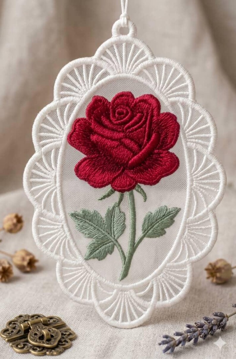 May include: An oval-shaped white lace ornament featuring a detailed embroidered red rose with green leaves and a stem. The lace border has a scalloped fan-like pattern, and a small white loop at the top allows for hanging this decorative piece.