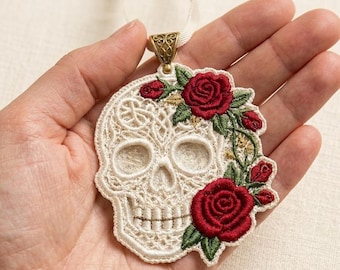 May include: An embroidered skull ornament in cream thread with intricate lace-like patterns. The skull is adorned with vibrant red roses and green leaves on the right side. It features a bronze-colored metal bail at the top with a cream ribbon.