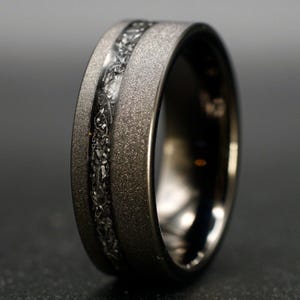 Könnte beinhalten: Ein dunkelgrauer Ring mit einer texturierten, sandgestrahlten Oberfläche an den Außenkanten und einer mittigen Einlage mit einem gezackten, metallisch silberfarbenen Fragmentmuster. Die Innenseite des Rings ist glatt, poliert und dunkel.