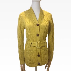 Puede incluir: Cárdigan de punto trenzado amarillo mostaza con cuello en V, mangas largas y cinturón a juego. El suéter tiene botones marrones y dos bolsillos delanteros. El cárdigan se exhibe en un maniquí.