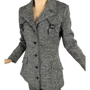 May include: A black and white herringbone pattern suit, consisting of a blazer and matching pants. The blazer has a notched collar, button closure, and decorative buckles. The suit is displayed on a mannequin.