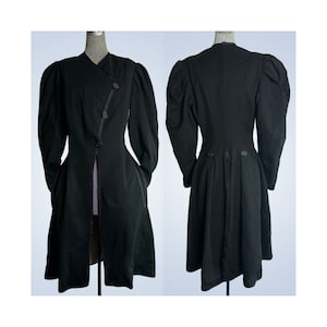 Puede incluir: Abrigo negro vintage con un cierre de botones asimétrico único y mangas abullonadas espectaculares. El abrigo presenta un diseño largo y fluido con un ajuste a medida, mostrando un estilo clásico y elegante. El abrigo se exhibe en un maniquí.