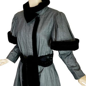 Puede incluir: Un abrigo gris de rayas diplomáticas con cuello alto de terciopelo negro, puños de terciopelo negro en las mangas abullonadas y un cinturón ancho de terciopelo negro para anudar en la cintura. La prenda tiene una silueta estructurada con mangas largas.
