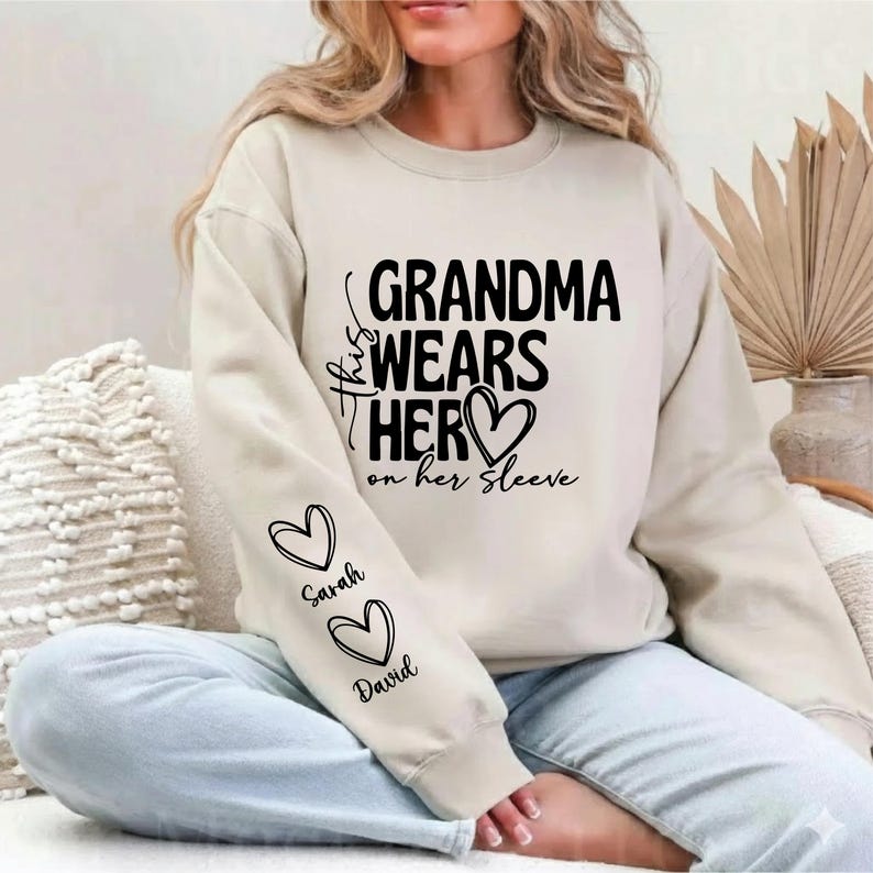May include: Beige sweatshirt with the text "Grandma Wears Her on Her Sleeve" in black. The sleeve features two heart graphics with the names "Sarah" and "David" below them. The sweatshirt is a casual style.