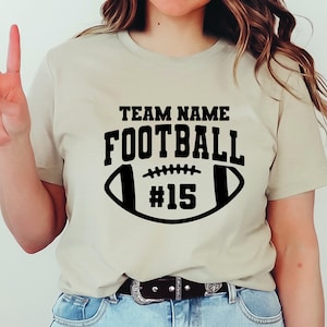 May include: A beige t-shirt with a black football graphic. The graphic includes the words "TEAM NAME" above "FOOTBALL" and the number "15" inside the football shape. The shirt is made of a soft material.