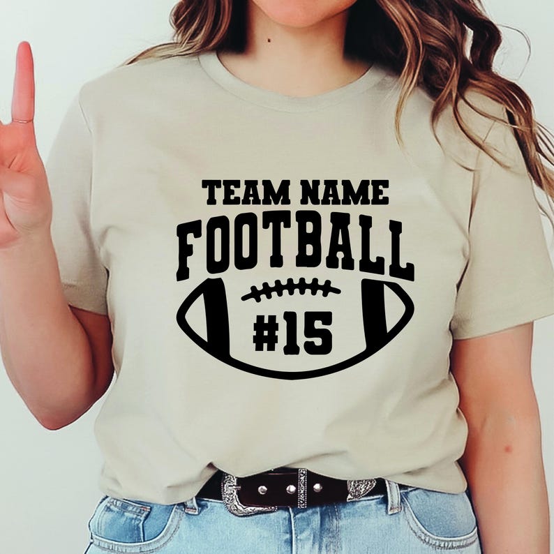 May include: A beige t-shirt with a black football graphic. The graphic includes the words "TEAM NAME" above "FOOTBALL" and the number "15" inside the football shape. The shirt is made of a soft material.