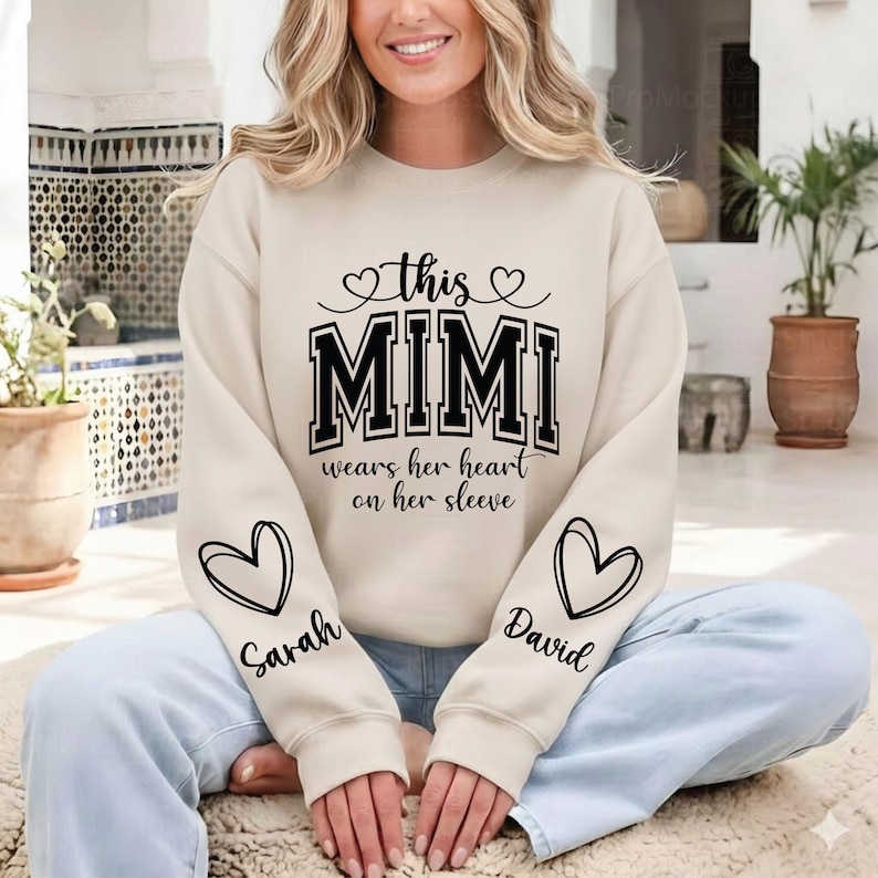 May include: Cream-colored sweatshirt with the text "this MIMI wears her heart on her sleeve" in black. The sleeves feature heart designs with the names "Sarah" and "David" in script. A cozy and personalized gift.