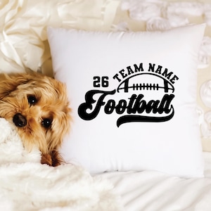 May include: White square pillow with a black football design. The design includes the number 26, the words "TEAM NAME", and the word "Football". A small, fluffy, brown dog is partially visible on the left side of the pillow.