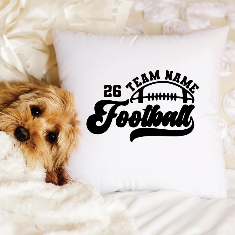 May include: White square pillow with a black football design. The design includes the number 26, the words "TEAM NAME", and the word "Football". A small, fluffy, brown dog is partially visible on the left side of the pillow.