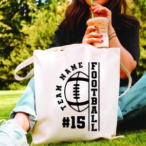 May include: Cream-colored tote bag with a black football graphic, the words "TEAM NAME" in a semi-circle, and "FOOTBALL" vertically. The number "15" is below the football. The bag has long handles.