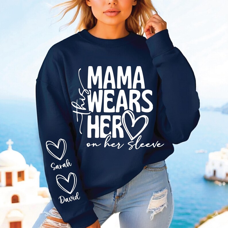 May include: Navy blue sweatshirt with the text "Mama Wears Her Hero on her Sleeve" in white. The sleeve features two white heart graphics with the names "Sarah" and "David" below them. The sweatshirt is a casual top.