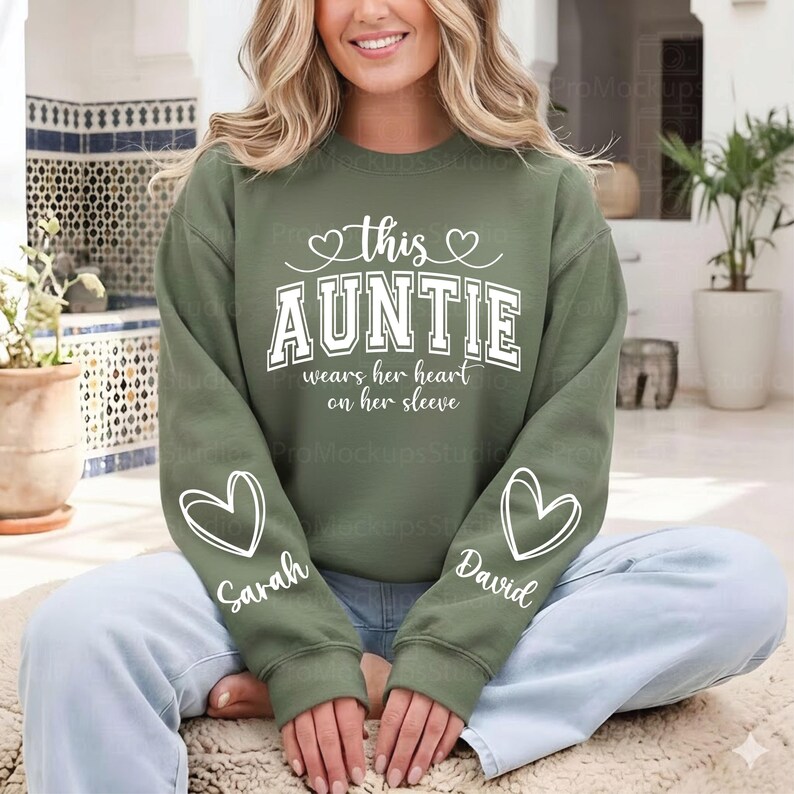 May include: Olive green sweatshirt with the text "this AUNTIE wears her heart on her sleeve" in white. The sleeves have white heart designs with the names "Sarah" and "David". The sweatshirt is a casual style.