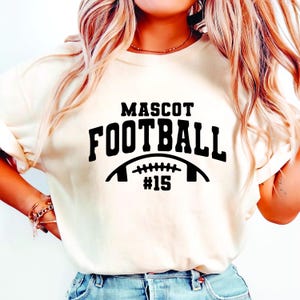 May include: Cream-colored t-shirt with the words "MASCOT FOOTBALL" in bold black letters. Below the text is a football graphic with the number "#15" in black. The shirt is a casual style.