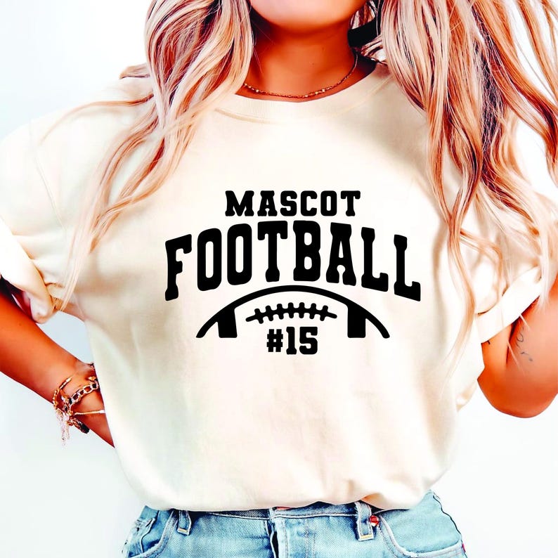 May include: Cream-colored t-shirt with the words "MASCOT FOOTBALL" in bold black letters. Below the text is a football graphic with the number "#15" in black. The shirt is a casual style.